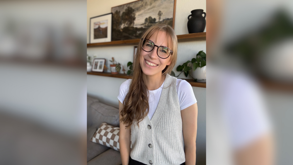 Photo of a woman with long dark blond hair smiling at the camera. The woman is wearing glasses and standing in front of shelves with office decorations on them.