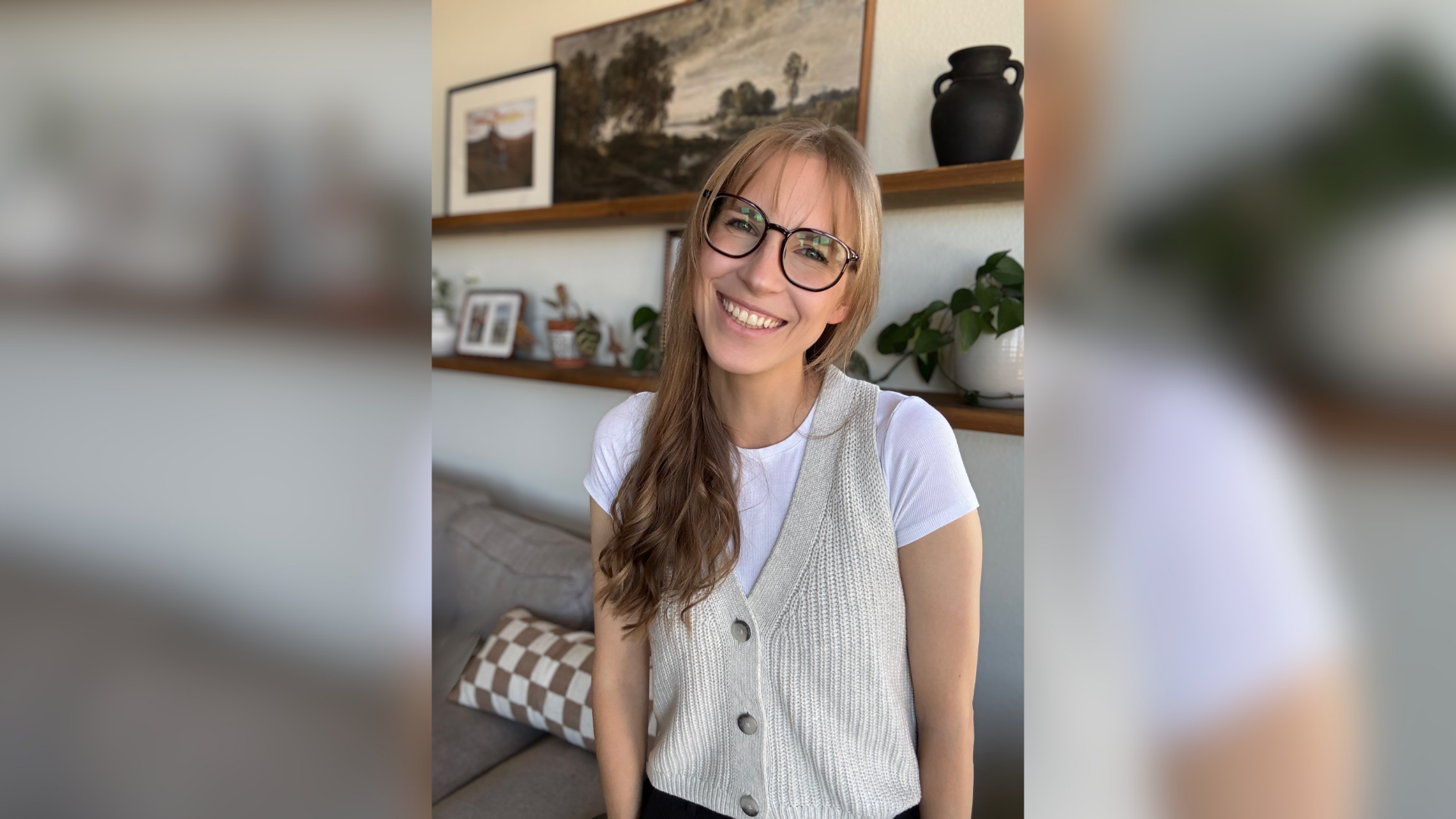 Photo of a woman with long dark blond hair smiling at the camera. The woman is wearing glasses and standing in front of shelves with office decorations on them.